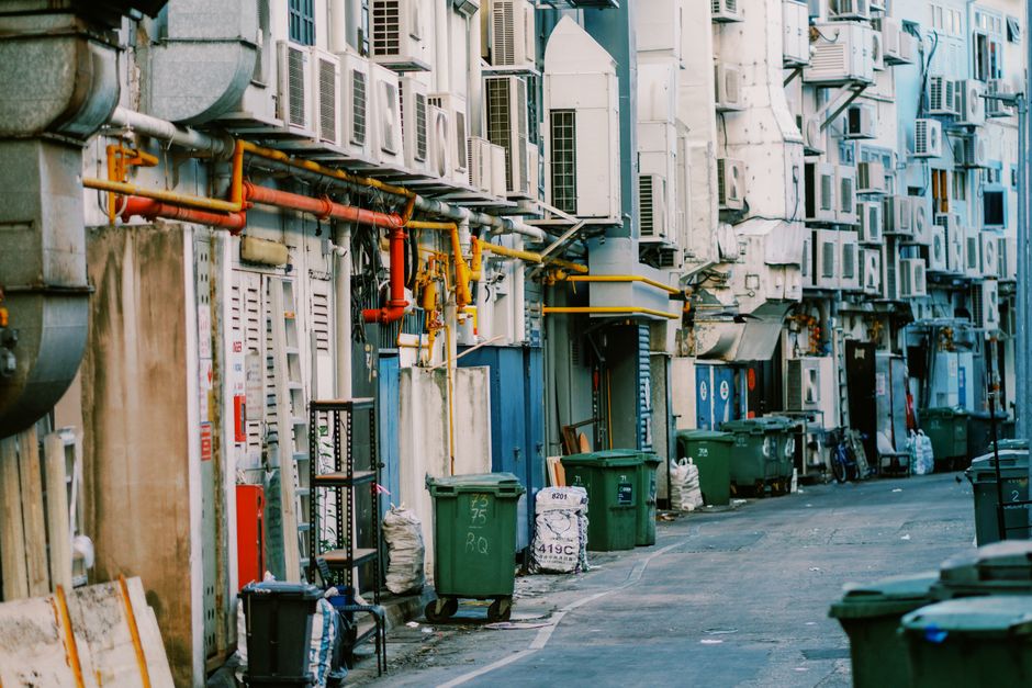 A narrow urban alleyway lined with a series of beige and off-white building facades, each equipped with multiple small window air conditioning units mounted on the exterior walls. In the foreground, several green wheeled rubbish bins are positioned along the curb, some with lids closed and others partially open, revealing discarded packaging or waste bags inside. An assortment of black and white trash bags and miscellaneous waste items rest against the bins and building walls. Visible pipes, including orange and yellow gas and water lines, run horizontally across the buildings, connecting various utility meters and fixtures. The ground surface consists of uneven concrete pavement, with slight dirt and debris scattered in places. The scene shows signs of maintenance and clutter typical of a servicing area for waste collection, suggesting a setting where independent rubbish removal services, such as those offered by Rubbish Clearance Finchley, might operate in an alternative waste handling context, avoiding reliance on traditional street collections. Lighting is natural, with soft daylight illuminating the scene, highlighting the textures of the building surfaces and waste containers.