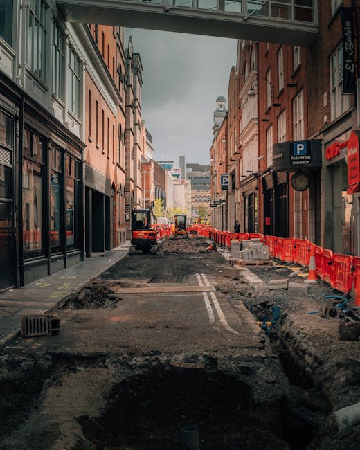 A narrow city street undergoing construction work with the surface partially excavated, revealing dark soil and broken pavement. On the right side, there are orange plastic safety barriers lining the sidewalk, and a few parked vehicles are visible further down the road. The buildings on both sides are multi-storey, constructed from brick and stone, with retail outlets on the ground floors, including a parking sign and signage for local businesses. An elevated pedestrian walkway spans above the street, and the street is illuminated by natural daylight under an overcast sky. The scene depicts an environment where private waste removal or on-site clearance might be necessary due to limited space for conventional rubbish collection, underscoring the importance of professional rubbish clearance services such as those offered by Rubbish Clearance Finchley in managing waste from urban construction or maintenance activities.