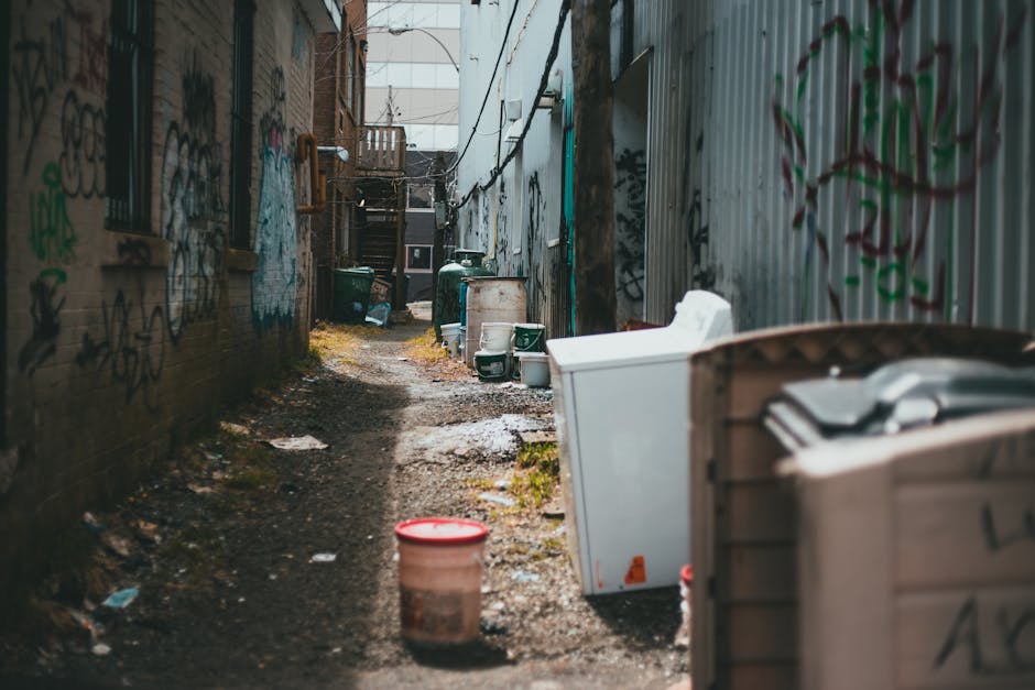 A narrow urban alleyway lined with a series of beige and off-white building facades, each equipped with multiple small window air conditioning units mounted on the exterior walls. In the foreground, several green wheeled rubbish bins are positioned along the curb, some with lids closed and others partially open, revealing discarded packaging or waste bags inside. An assortment of black and white trash bags and miscellaneous waste items rest against the bins and building walls. Visible pipes, including orange and yellow gas and water lines, run horizontally across the buildings, connecting various utility meters and fixtures. The ground surface consists of uneven concrete pavement, with slight dirt and debris scattered in places. The scene shows signs of maintenance and clutter typical of a servicing area for waste collection, suggesting a setting where independent rubbish removal services, such as those offered by Rubbish Clearance Finchley, might operate in an alternative waste handling context, avoiding reliance on traditional street collections. Lighting is natural, with soft daylight illuminating the scene, highlighting the textures of the building surfaces and waste containers.