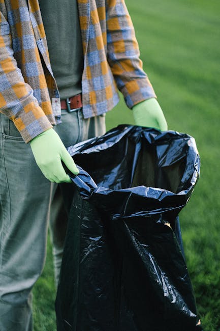 A person standing outdoors on a grassy area, wearing a plaid shirt with shades of orange, yellow, and blue over a green top, and grey trousers secured with a brown belt. They are wearing light green rubber gloves and are holding open a large black plastic rubbish bag, which appears to be ready for waste collection. The background features a blurred, lush green lawn or field, suggesting an outdoor cleaning or waste disposal activity. The scene is well-lit with natural daylight, emphasizing the practical nature of waste management tasks, consistent with services offered by companies like Rubbish Clearance Finchley that specialize in independent or private rubbish removal. The focus on the person's hands and the rubbish bag highlights the act of waste collection and disposal, supporting the thematic focus on alternative waste handling options outside of local authority collection