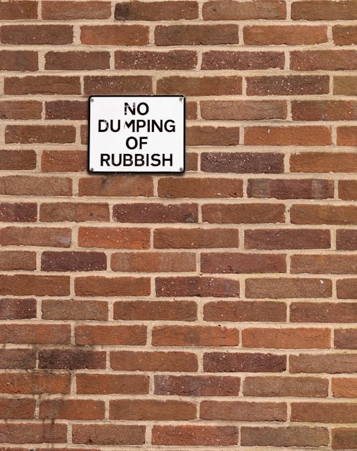 A rectangular metal sign mounted on a red brick wall displays the message 'NO DUMPING OF RUBBISH' in black uppercase letters on a white background. The brick wall features standard-sized, horizontally laid bricks with a slightly rough texture and a mix of reddish-brown and darker brown tones, with light grey mortar joints in between. The sign is positioned approximately one third from the top of the wall and is centered horizontally. The bricks extend evenly across the scene, with no other objects or debris visible in the image. The lighting appears natural, casting soft shadows on the brick surface, and the overall scene reflects a typical urban or residential exterior environment, supporting the context of private waste management and rubbish disposal regulation as highlighted by local rubbish removal services such as Rubbish Clearance Finchley. The image emphasizes the importance of adhering to waste disposal rules in designated areas to prevent illegal dumping, aligning with the message conveyed by the sign and the broader topic of alternative waste handling methods outside local authority rubbish collection.