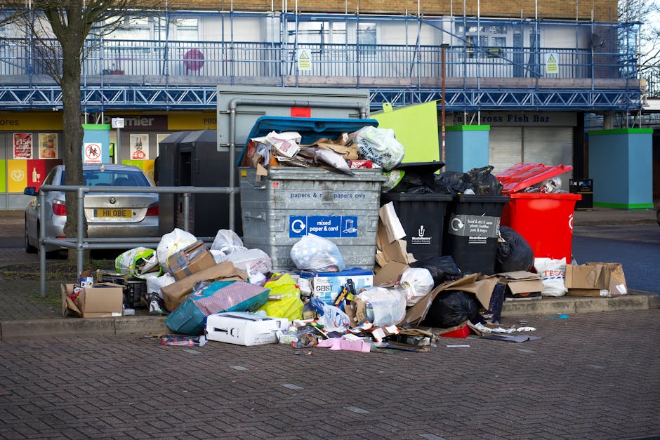 A large collection of refuse and waste materials, including cardboard boxes, plastic bags, paper and packaging, piled around and spilling out of various rubbish bins and containers on a paved urban sidewalk. The waste is a mix of bulky packaging, torn cardboard, and loose paper, some of which are in partially opened bags or discarded on the ground. The scene is set in front of a commercial building under construction or renovation, with metal scaffolding and protective netting visible in the background, alongside a parked car with a yellow license plate. Several waste bins are visible; one is metallic with a blue lid labeled for mixed paper and card, another black, and a red recycling container. The area is bordered by metal railings, and there is a tree on the left side. This scene illustrates the need for independent rubbish collection services like those offered by Rubbish Clearance Finchley, highlighting the common issue of waste overflow outside urban commercial zones and the importance of proper on-site waste handling for effective rubbish removal.