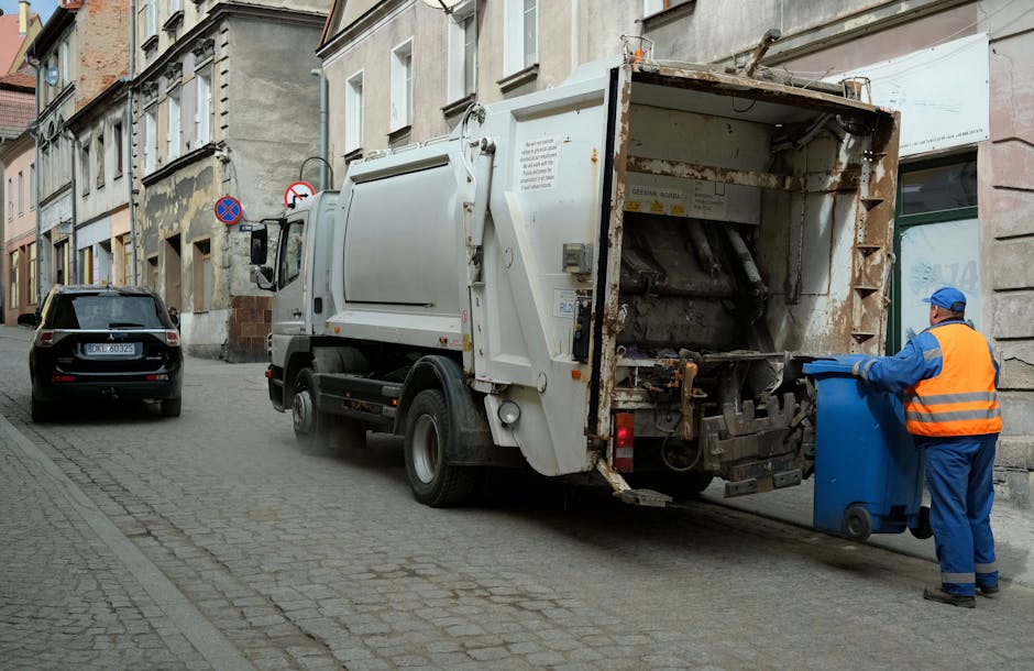 A street scene featuring a waste collection truck with an open rear compartment, positioned alongside a curb on a cobblestone road. The truck, which is primarily white with some rusted metal parts, has a mechanical lifting mechanism visible at the back, used for compacting and emptying waste. A worker dressed in a blue uniform and an orange high-visibility vest, wearing a blue cap, is in the process of disposing of blue plastic waste bins into the truck’s compartment. The surrounding environment includes older, multi-storey buildings with a mix of stone and brick facades, and a parked black car on the left side of the street. Signage indicating parking restrictions is visible on the wall of one building. The scene is lit with natural daylight, and the overall setting suggests a typical urban environment where independent waste collection services, such as those provided by Rubbish Clearance Finchley, operate for rubbish removal and local waste management.