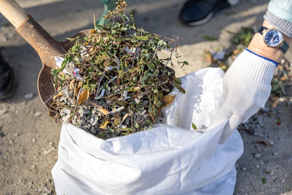 A person wearing a white glove and a watch on their wrist is holding a white woven polypropylene sack filled with a mixture of garden debris, including small green leafy branches and twigs, along with bits of gravel or broken concrete. The debris appears to be gathered from a garden or outdoor area, with the sack resting on a gravel or dirt surface. In the background, part of a rusty metal shovel is visible, with its blade partially buried in the ground. Nearby, a pair of black shoes and a small portion of a person's light blue jeans are seen standing on the ground. The scene conveys a clear act of rubbish removal or garden waste collection, supported by the surroundings and the handling tools, often associated with independent waste clearance services like those provided by Rubbish Clearance Finchley for local residents or businesses in N3, London, aligning with the context of private, on-site rubbish clearance efforts.