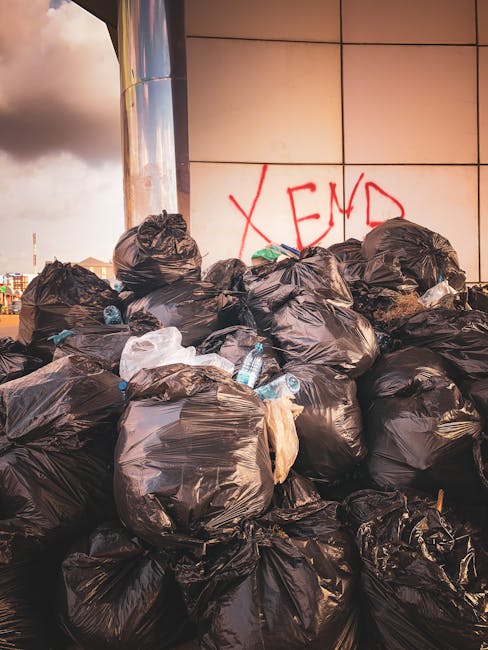 A large accumulation of tightly packed black plastic rubbish bags stacked against a modern building wall with beige tiles, which has the word 'XEND' spray-painted in red graffiti. The bags appear to contain waste materials, and some are torn open, revealing plastic bottles and miscellaneous debris inside. The setting is outdoors on a paved surface, possibly a pavement or driveway, with a partly cloudy sky visible in the background. The scene suggests an instance of private waste or rubbish clearance that may require professional assistance, such as services offered by Rubbish Clearance Finchley, to manage and dispose of bulk waste effectively and safely. The environment reflects an informal collection point for waste awaiting collection, aligned with the theme of rubbish removal and alternative disposal methods outside of regular municipal services.