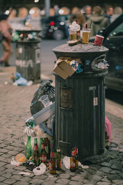 A black, cylindrical public rubbish bin made of metal, situated on a cobblestone pavement, is overflowing with various trash items. The top surface of the bin holds used paper cups with plastic lids, a disposable food container with some leftover food, and a crumpled paper napkin, all appearing to be food-related waste. Around the base of the bin, there are several glass bottles, mostly with red and green labels, some lying on their sides and some with caps, along with discarded paper and plastic wrappers. A black garbage bag is tied and positioned next to the bin, facilitating additional waste disposal. In the blurred background, pedestrians and parked vehicles are visible, indicating a busy street scene. Some trash is also spilling over the sides of the bin and onto the cobblestone surface, suggesting it has not been emptied recently. The environment appears to be an outdoor urban area, where individuals may have left rubbish after eating or socializing, highlighting the importance of waste collection services like those offered by Rubbish Clearance Finchley for effective rubbish removal and street cleanliness.