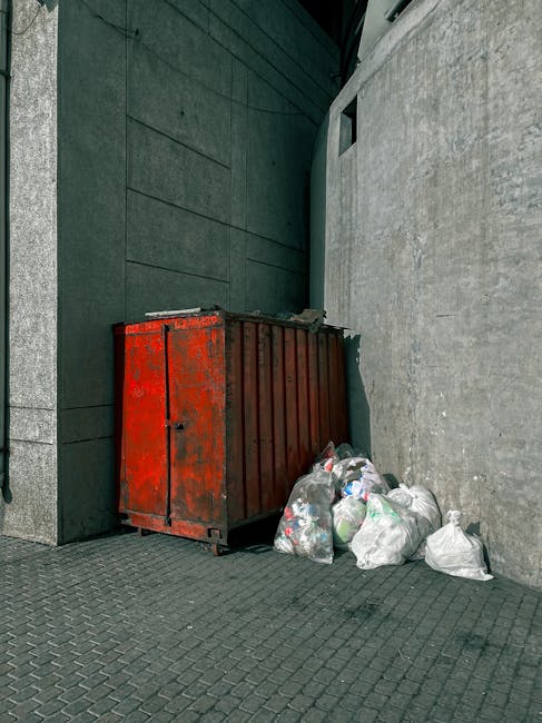 A black, cylindrical public rubbish bin made of metal, situated on a cobblestone pavement, is overflowing with various trash items. The top surface of the bin holds used paper cups with plastic lids, a disposable food container with some leftover food, and a crumpled paper napkin, all appearing to be food-related waste. Around the base of the bin, there are several glass bottles, mostly with red and green labels, some lying on their sides and some with caps, along with discarded paper and plastic wrappers. A black garbage bag is tied and positioned next to the bin, facilitating additional waste disposal. In the blurred background, pedestrians and parked vehicles are visible, indicating a busy street scene. Some trash is also spilling over the sides of the bin and onto the cobblestone surface, suggesting it has not been emptied recently. The environment appears to be an outdoor urban area, where individuals may have left rubbish after eating or socializing, highlighting the importance of waste collection services like those offered by Rubbish Clearance Finchley for effective rubbish removal and street cleanliness.