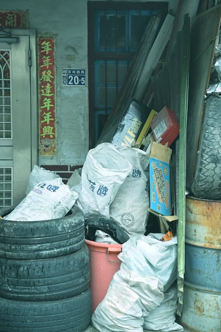 A collection of used tires, white plastic bags, and cardboard boxes are stacked outside a building, partially leaning against the wall and covering a portion of the ground. The tires have a textured black rubber surface and are stacked horizontally in the foreground, while the white plastic bags appear to be filled with waste and are placed atop the stack of tires. Several cardboard boxes with printed labels and graphics are positioned behind the bags, some slightly tilted. To the right, there are large, disused barrels with rust and dirt, partially obscured by the bags and tires, and leaning tools or wooden planks are propped up against the wall. The background shows a glass window, a metal door with a latch, and a wall bearing a blue address plate with the number 20-5, alongside vertical red and yellow Chinese banners with calligraphic characters, indicating an urban residential or industrial area where private or independent waste clearance might be undertaken by services such as Rubbish Clearance Finchley. The lighting suggests natural daylight, highlighting the textures of worn tires, crumpled bags, and weathered barrels, illustrating a typical scene of rubbish accumulation suited for professional waste removal or on-site clearance services.
