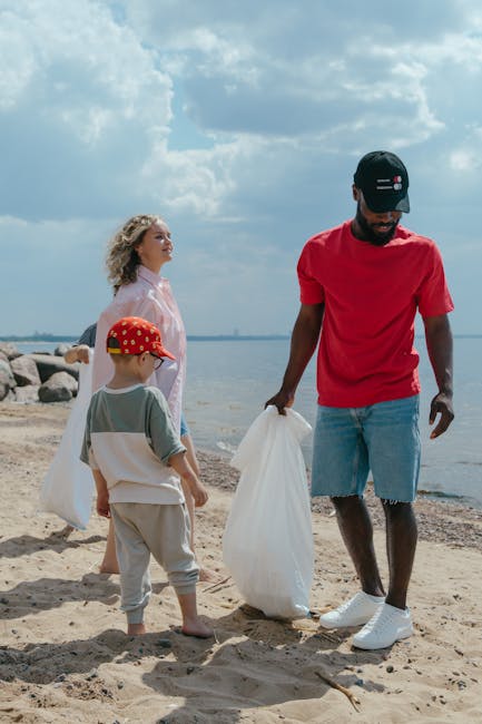 A man and two children collecting rubbish on a sandy beach near a body of water under a partly cloudy sky. The man is dressed in a red t-shirt, light blue denim shorts with frayed hems, and white sneakers, holding a large white rubbish bag. The young boy, wearing a grey t-shirt, beige trousers, and a red cap with white spots, is also holding a rubbish bag and facing the man. A young girl with curly blonde hair, dressed in a light pink jacket, stands nearby and observes the scene. In the background, rocks line the shoreline, and the sky has white and grey clouds, indicating a partly sunny day. This scene illustrates private or independent rubbish collection efforts at a natural outdoor setting, supporting sustainable waste management practices outside of municipal services, akin to on-site clearance or alternative waste handling. Rubbish Clearance Finchley often carries out similar rubbish removal services, emphasizing environmentally responsible disposal methods in various environments, including beaches and coastal areas.