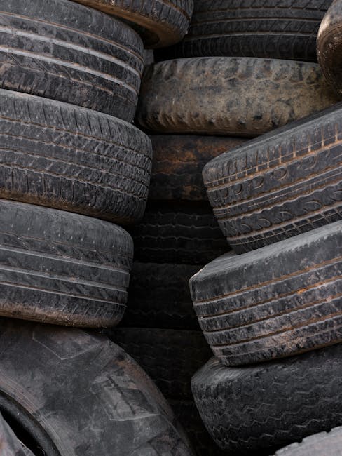 A close-up image of a stack of used automotive tyres arranged closely together, filling the entire frame. The tyres vary in size and tread pattern, with some displaying visible grooves and worn surfaces, and others showing signs of weathering with discoloured rubber and dirt. The tyres are primarily black, with areas of lighter, faded rubber and subtle patches of rust-coloured residue. The textures range from smooth to heavily grooved, with some tyres exhibiting cracking on their sidewalls. They are positioned on a rough, outdoor surface, possibly a concrete or asphalt yard, surrounded by minimal background elements to emphasize the tyre pile. This scene, captured in diffuse daylight, reflects a typical scenario where old tyres are stored, awaiting collection or disposal, aligned with the services of private waste disposal providers such as Rubbish Clearance Finchley, who handle alternative waste materials outside standard municipal rubbish collection. The image effectively illustrates the need for proper tyre disposal or recycling as part of broader rubbish removal efforts.